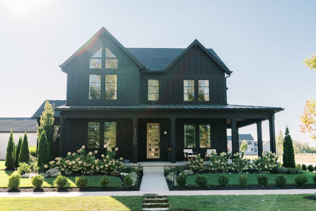A modern two-story black house with large windows, a covered front porch, and a well-maintained garden with shrubs and flowers.