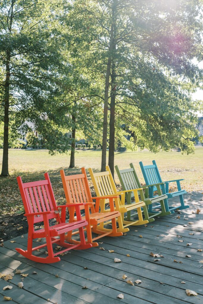 Six colorful rocking chairs—red, orange, yellow, green, blue, and teal—lined up on a wooden deck outdoors, with trees and sunlight in the background.