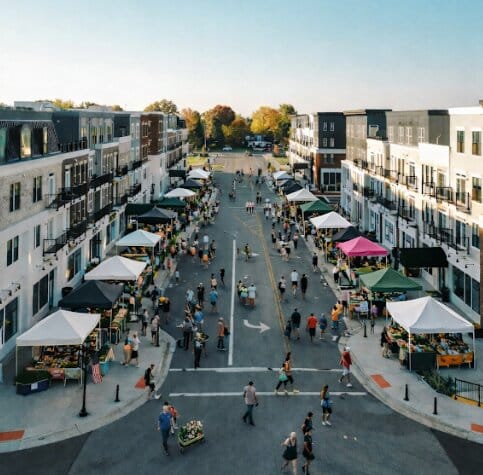 Aerial view of a bustling farmers market lined with vendor tents and people walking between modern apartment buildings on a clear day.