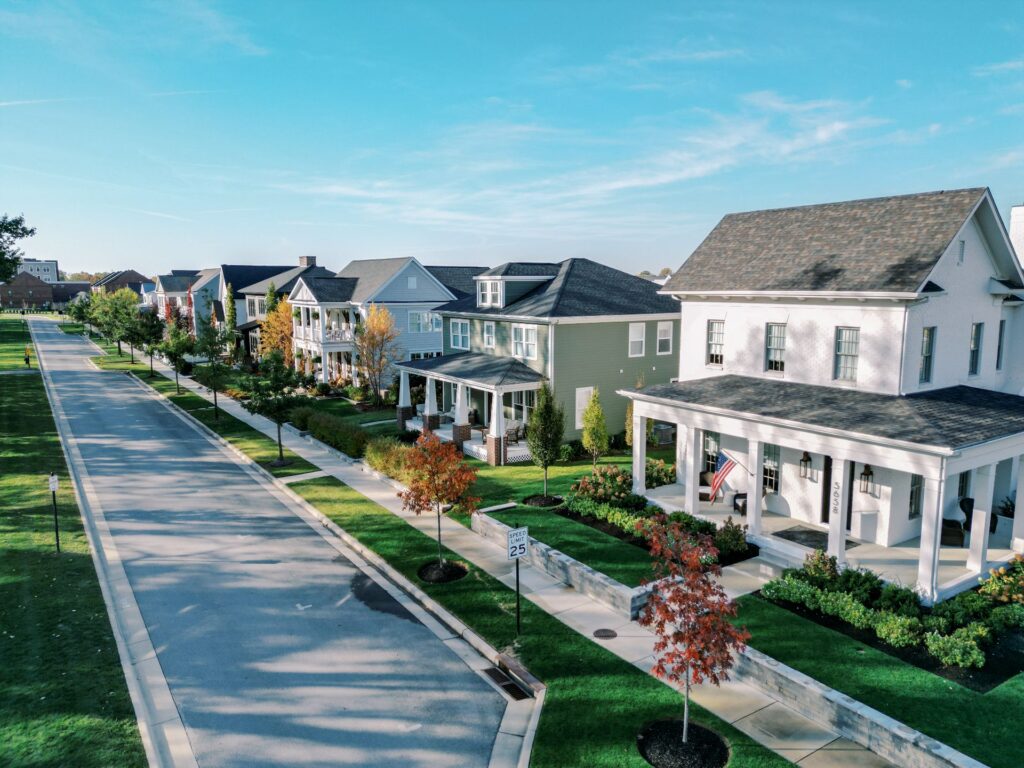 A row of modern suburban houses with front porches lines a clean, tree-lined street under a clear blue sky.
