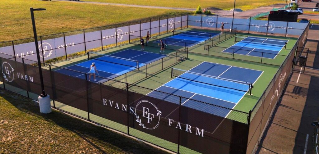 Aerial view of four outdoor pickleball courts at Evans Farm, where several people are playing and others stand nearby. Discover Evans Farm Location - Ideal Living in Lewis Center, OH.