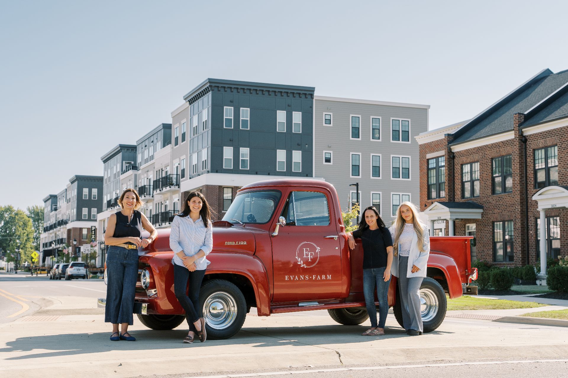 Four women, Connect and Thrive: Evans Farm Residents, stand in front of a red Evans Farm pickup truck parked on a street lined with modern apartment buildings and townhouses in the background.