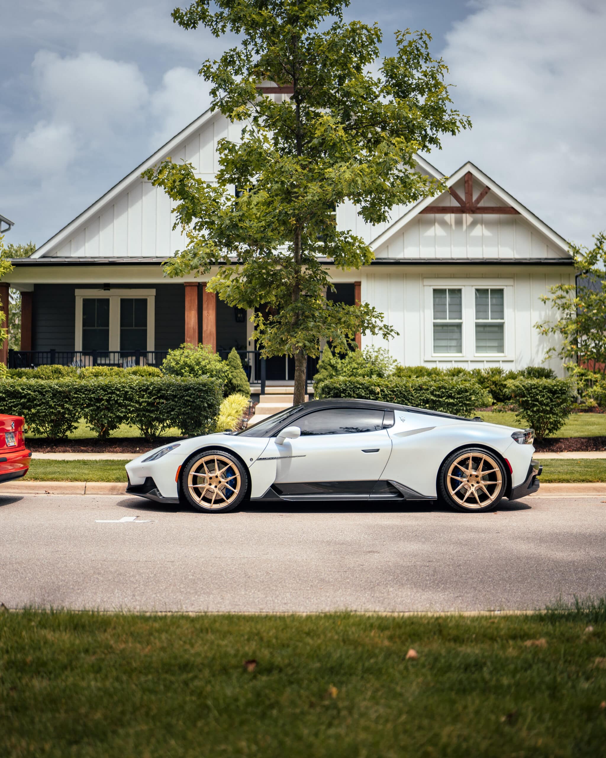 A white sports car with gold wheels is parked on the street in front of a modern suburban house with a front porch and green landscaping.