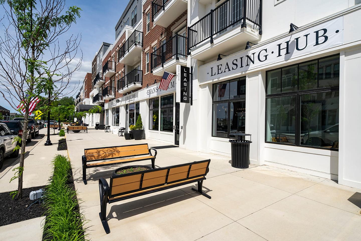 Modern apartment building with leasing office, benches, and young trees along a clean sidewalk on a sunny day. Contact Evans Farm for leasing information.