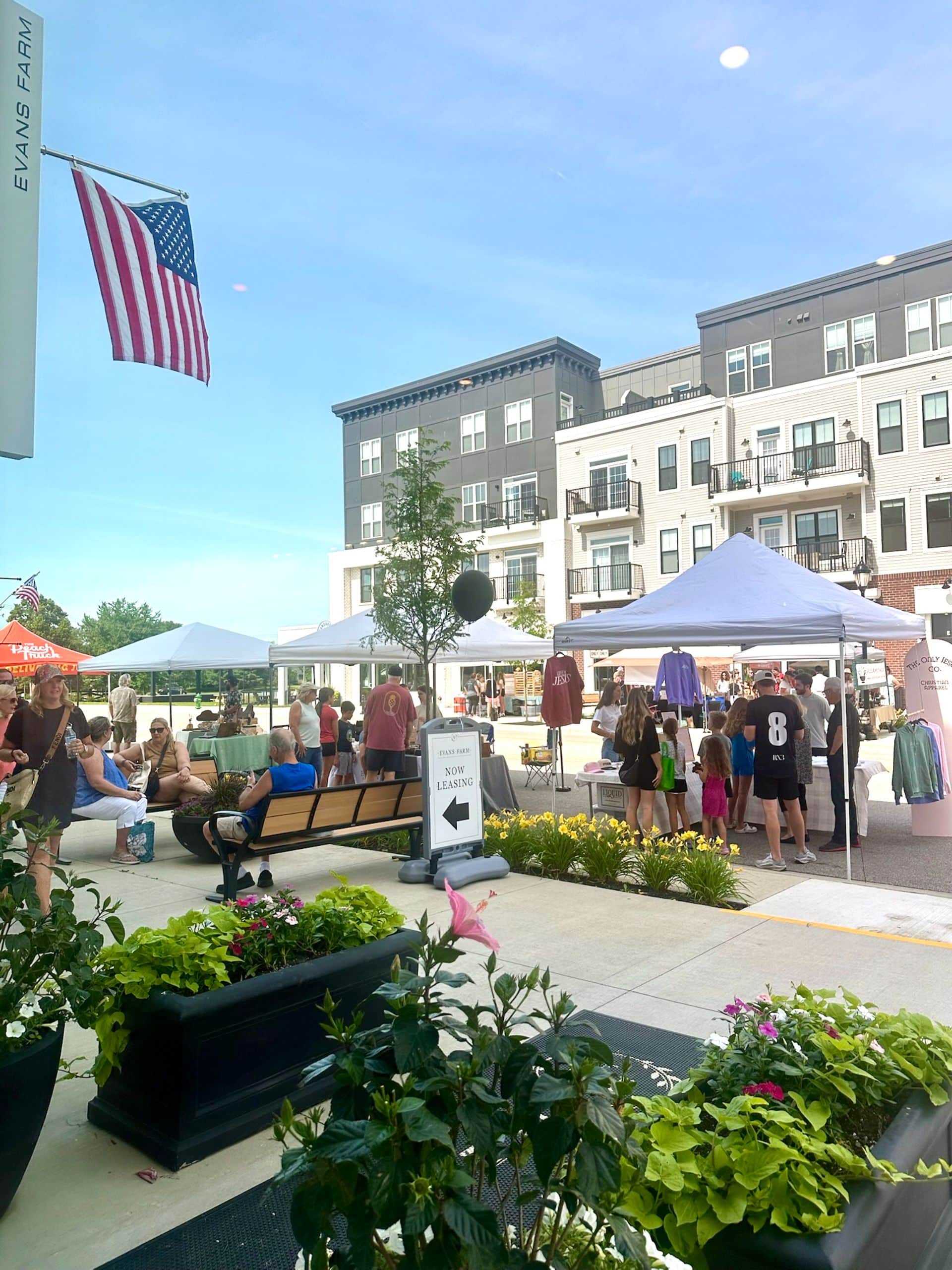 People gather around vendor tents at an outdoor market in a modern plaza, with an American flag and apartment buildings in the background. Benches and planters are visible in the foreground.