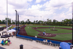 A youth baseball game is being played on an artificial turf field, with spectators seated in bleachers and players positioned on the field under a partly cloudy sky.