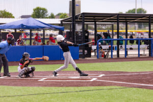 A youth baseball player in a black jersey swings at a pitch while the catcher and umpire look on during a game on an artificial turf field.