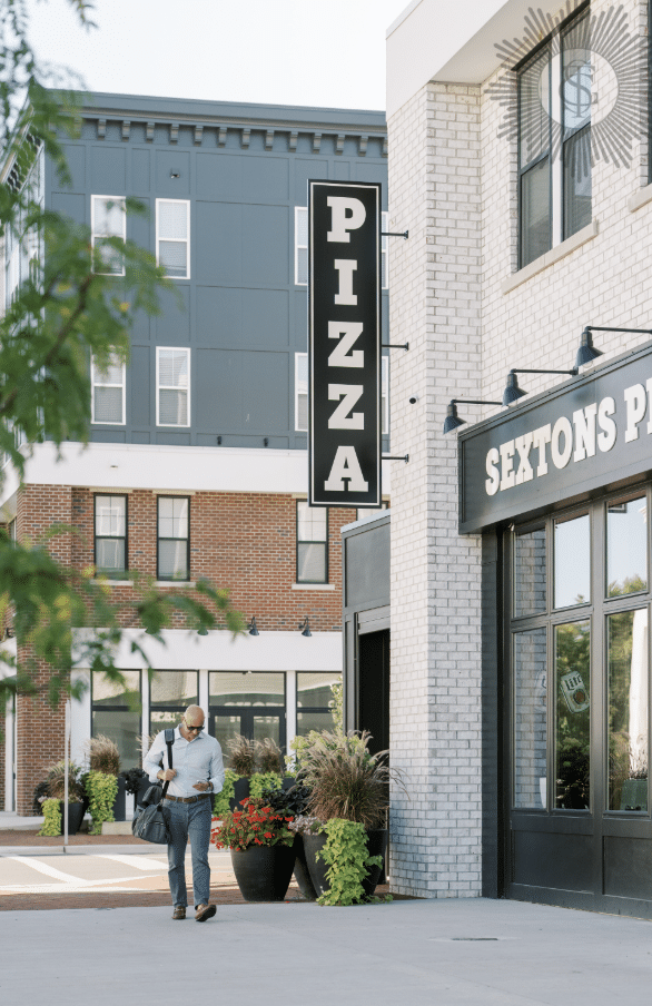 A person walks past a brick building with a large vertical "PIZZA" sign and a storefront labeled "SEXTONS PIZZA" in a modern urban area—just minutes from Discover Evans Farm Location, ideal living in Lewis Center, OH.