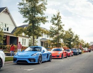 A row of sports cars, including a blue one in front, is parked along a suburban street with houses and trees; a few people walk nearby.