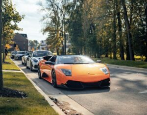 An orange Lamborghini convertible is parked on a tree-lined street with several other cars behind it on a sunny day.
