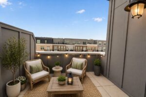 A small rooftop patio with two cushioned chairs, a table with a lantern, potted plants, string lights, and neighboring buildings in the background under a clear sky.