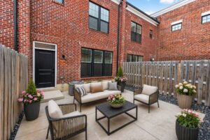 Enclosed patio with outdoor seating, potted flowers, and a black door, surrounded by a wooden fence and brick walls of the house.