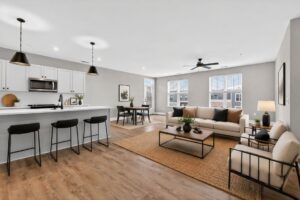 Modern open-concept living area with kitchen, dining, and lounge space; neutral tones, wood flooring, bar stools, sofa, chairs, and large windows providing natural light.