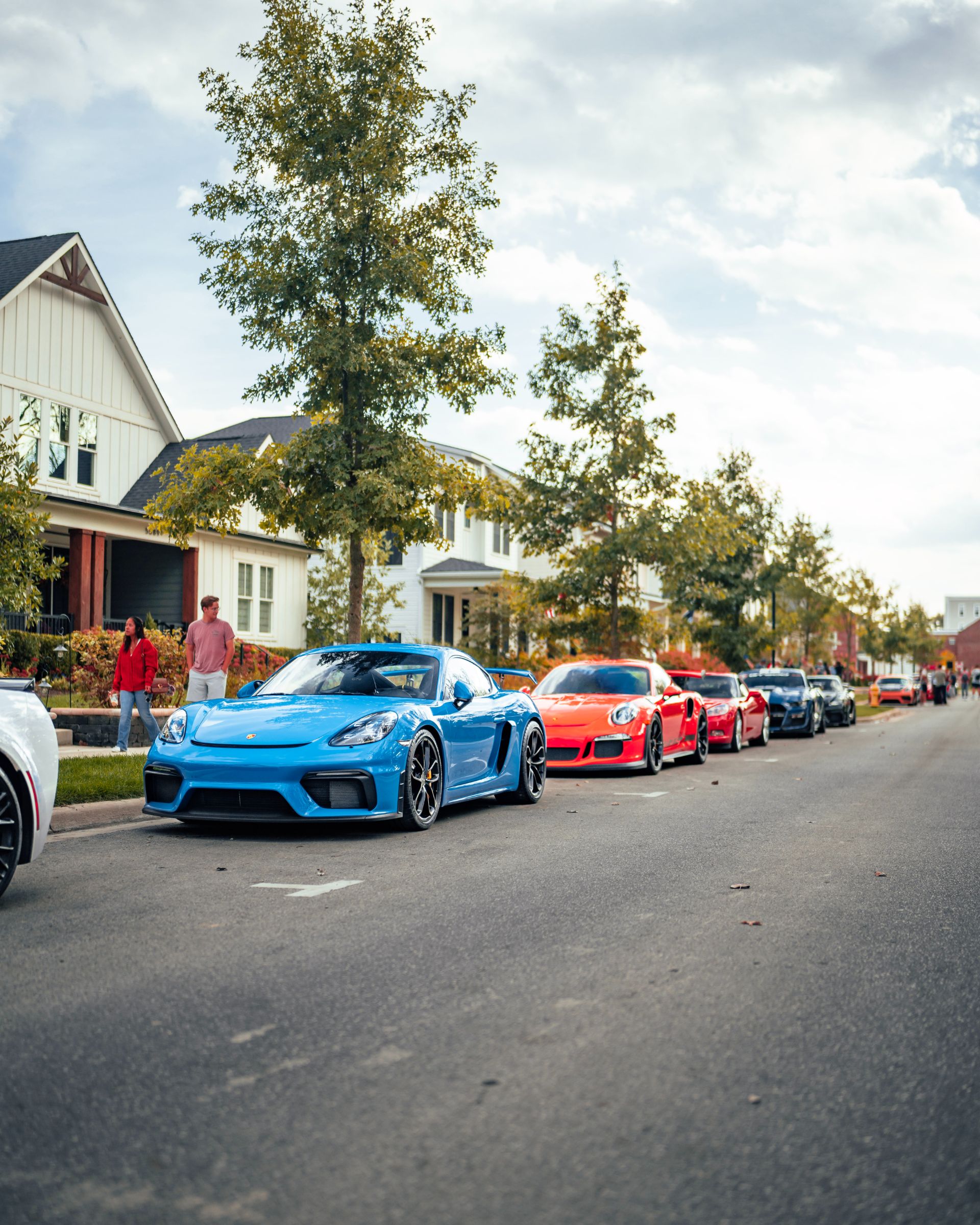 A row of colorful sports cars, including a blue and a red car, are parked along a residential street lined with trees and houses under a partly cloudy sky.
