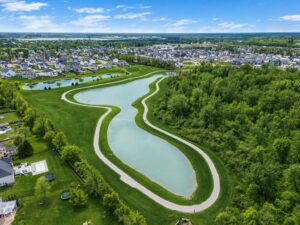 A winding pond bordered by a walking path runs alongside a residential neighborhood and wooded area under a partly cloudy sky.