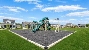 A playground with slides, swings, and climbing structures sits on mulch in the center of a grassy field, surrounded by suburban houses under a blue sky.