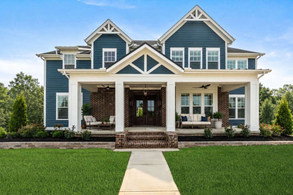 Two-story blue house with white trim, large front porch, brick steps, manicured lawn, and landscaping; clear blue sky in the background—Explore Homeownership in Lewis Center with this charming property.