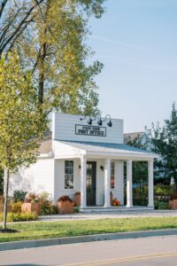 A small white building labeled "Evans Farm Post Office" has a front porch, potted plants, autumn decorations, and is surrounded by trees and greenery.