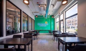 A modern café with black tables and chairs, large windows, concrete floor, and a green wall featuring a neon sign that reads "Sunny Side Up Café.