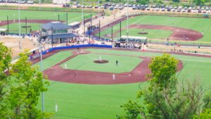 Aerial view of a baseball field with players in position, adjacent to another field, with spectators and parked cars in the background.