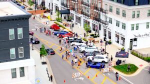 Aerial view of a street event with parked cars, pedestrians, and barricades in a modern urban shopping area. People gather around vehicles while others walk along the sidewalks.