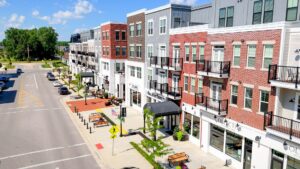 Street-level view of a modern mixed-use building with shops on the ground floor, residential units above, parked cars, and a clear blue sky.