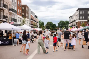 People walk among vendor tents at an outdoor street market lined with apartment buildings on a cloudy day.