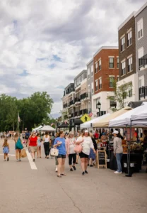People walk along a closed street lined with vendor tents and mid-rise buildings during an outdoor market on a cloudy day.