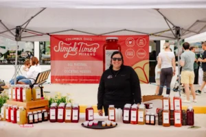 A woman stands behind a table displaying bottles of cocktail mixers at an outdoor market stall with a "Simple Times Mixers" banner.
