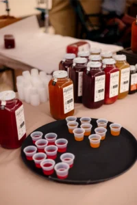 Bottles of juice with labeled tags are displayed behind a tray of small sample cups filled with red and orange liquid on a table.