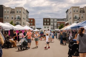 People walk and shop at an outdoor street market lined with vendor tents, with modern apartment buildings in the background under a cloudy sky.
