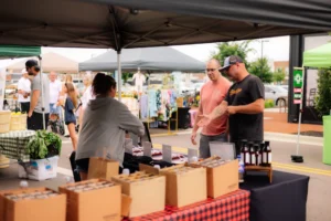 People interact at an outdoor market stall with boxes of goods, bottles, and other products displayed on tables under tents.