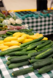 Yellow squash and green cucumbers are displayed on a checkered table at a market, with other vegetables visible in the background.