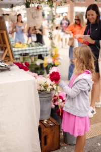 A young girl in a pink dress holds flowers at a market stall with buckets of flowers, while adults and other shoppers are in the background.