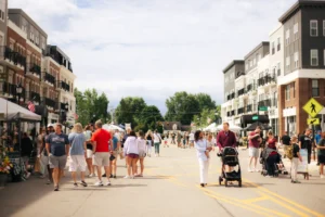 People walk along a closed street lined with tents and buildings during a daytime outdoor event or street fair.