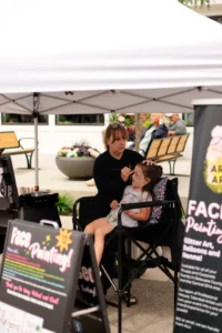 A woman paints a young girl’s face while she sits in a chair under a white tent at an outdoor face painting booth. Signs and art supplies are visible in the foreground.