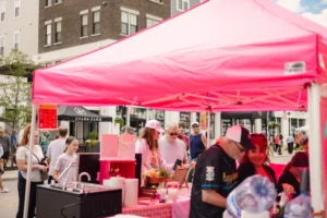 People gather under a bright pink canopy at an outdoor market or street fair, with various food and drink items displayed on a table in the foreground.