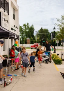 Several children play with scooters and toys on a sidewalk near a building, while two adults supervise and push a stroller. Balloons and outdoor decorations are visible.