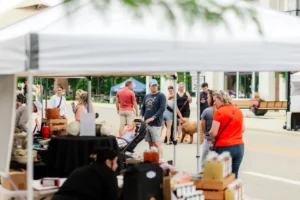 People browse stalls at an outdoor market on a city street, with various goods displayed and a few dogs and strollers visible among shoppers.