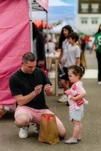 An adult kneels by a paper bag, holding an object, while a young child holding a pink stuffed toy stands nearby at an outdoor event.