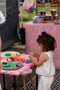 A young child stands at a small table covered with art supplies, focusing on a craft project at an outdoor event.