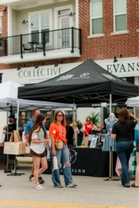 People walk past vendor tents at an outdoor market in front of a brick building with balconies and storefronts visible in the background.