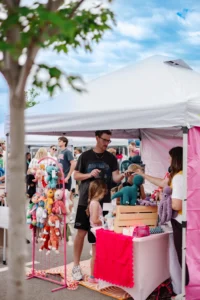 A man and child shop for handmade stuffed toys at an outdoor market stall with a pink and white tent, surrounded by other shoppers.