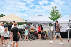 People walk and shop at an outdoor market with vendor tents under a blue sky. A bike is parked in front of a stall selling organic meats. Children and adults are visible in casual attire.