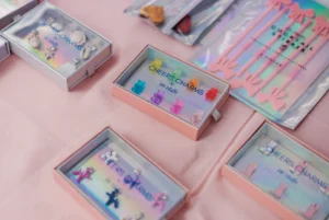 Several small boxes of colorful drink stirrers and charms are displayed on a pink tablecloth at a market or event.