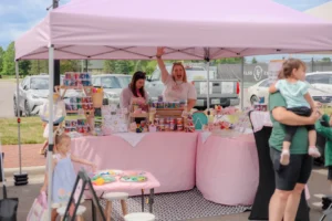 Two women stand at a booth under a pink canopy selling crafts and toys; one waves while children and adults walk nearby.