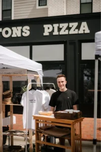 A person stands behind a small outdoor market stall selling shirts and accessories in front of a building with a pizza sign.