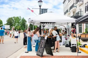 People browse goods and chat at outdoor vendor tents during a street market event on a sunny day, with several buildings and trees in the background.