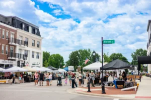 People browse vendor tents at an outdoor market on a city street under a partly cloudy sky. Buildings and a street sign for Market St are visible.