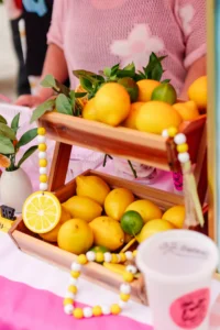 Two wooden trays filled with lemons, limes, and leaves are displayed on a pink table with a lemon-themed bead garland and a cup in the foreground.